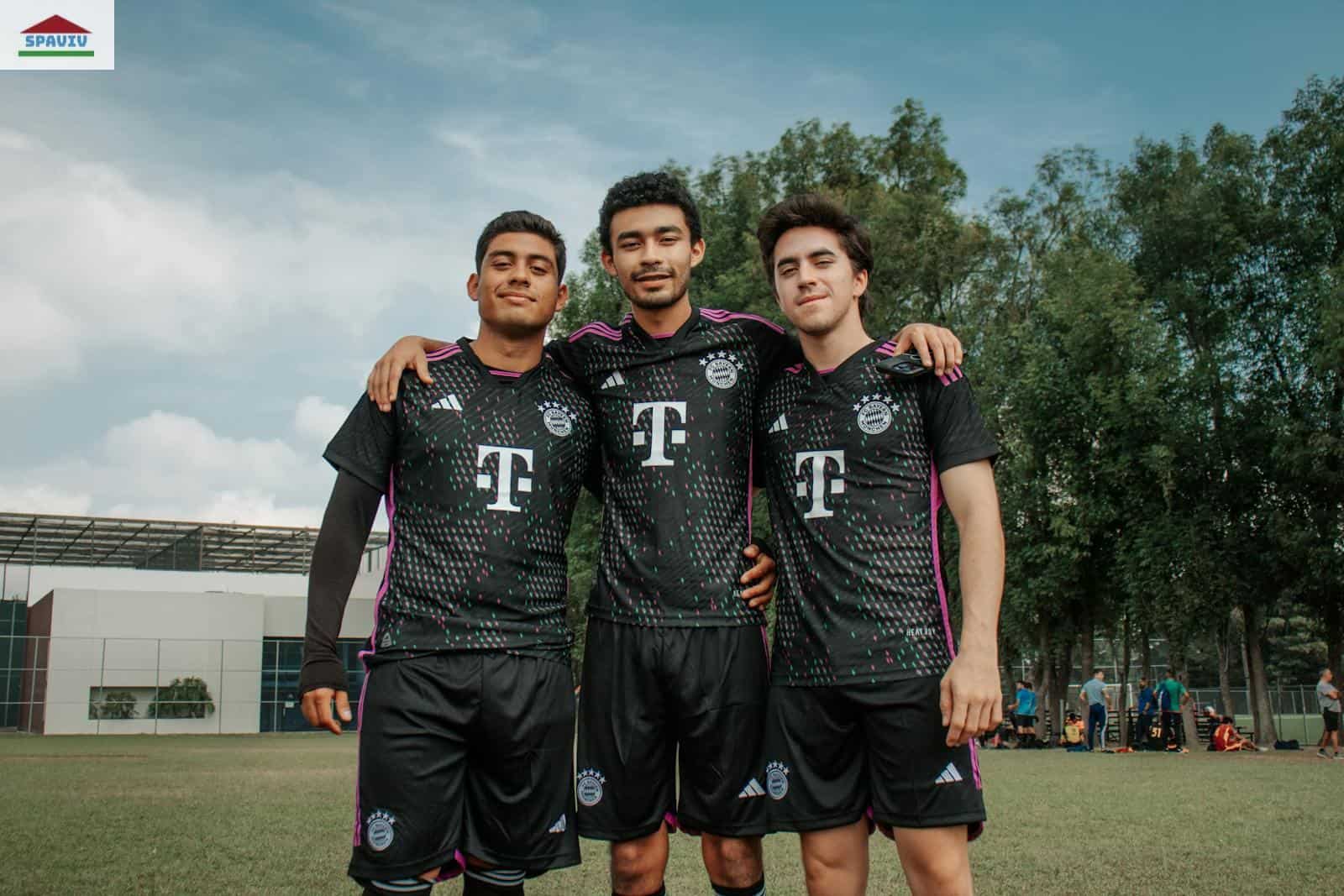 Three young adult soccer players in matching jerseys standing arm in arm outdoors.